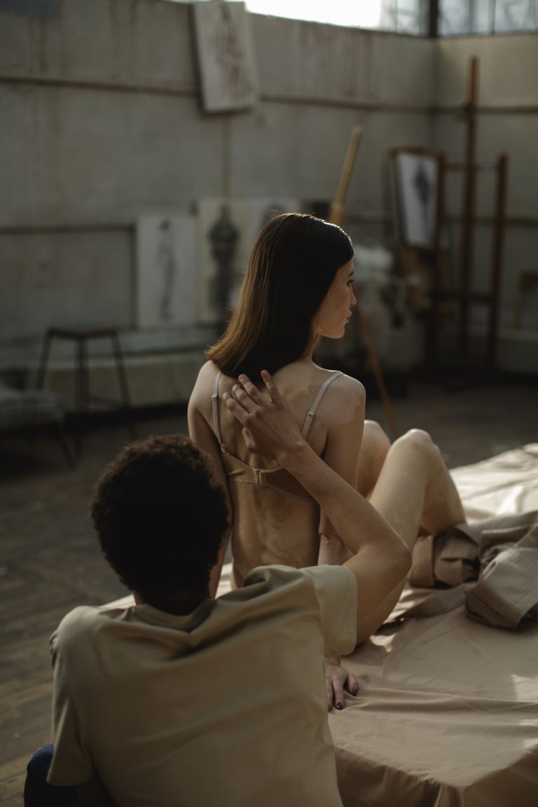 A model sits gracefully in an art studio while an artist observes closely.