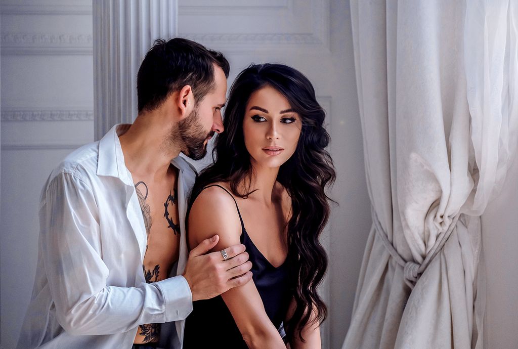 Calm young boyfriend in unbuttoned shirt hugging attractive woman wearing black silk nightgown while spending sunny morning together in light room