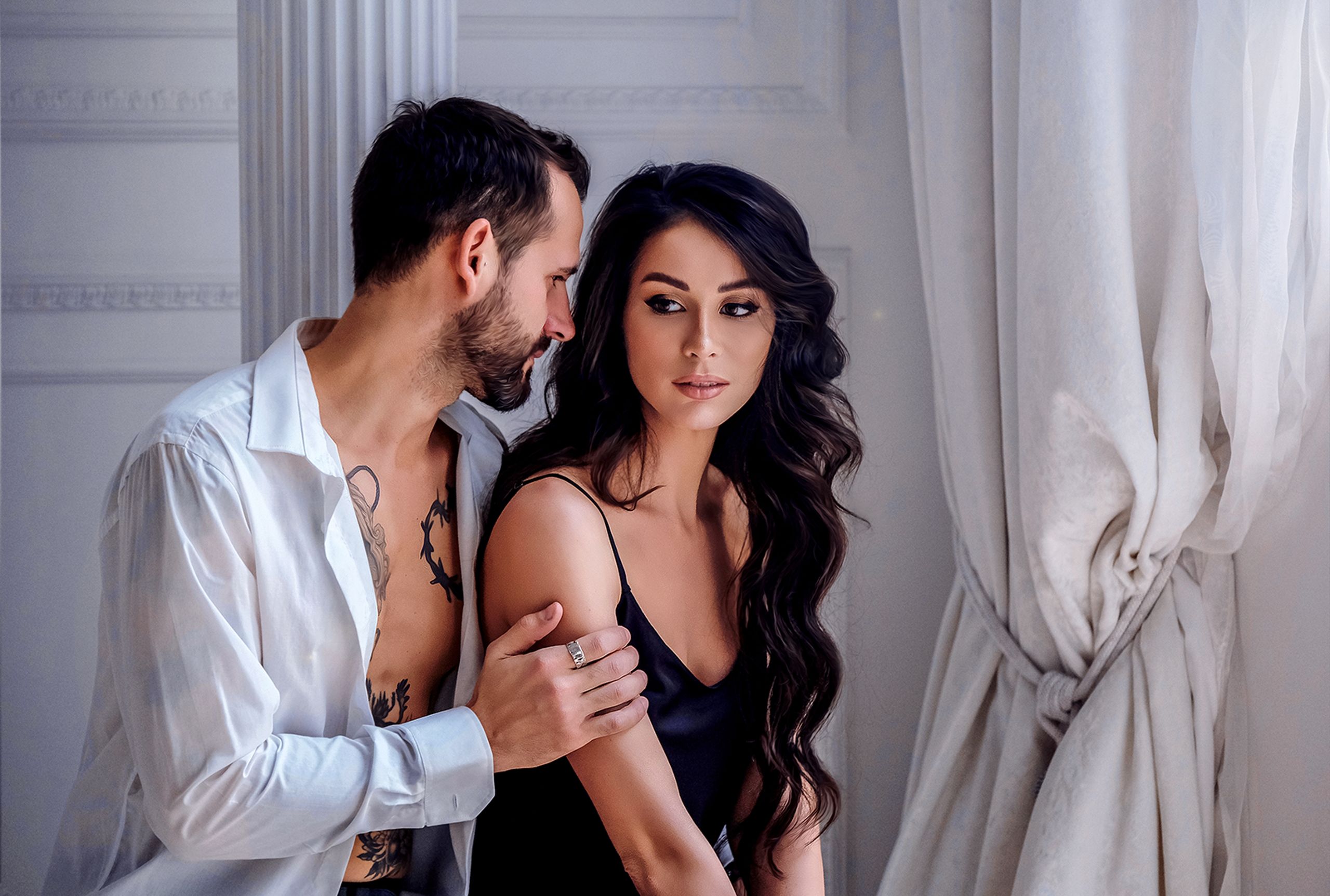Calm young boyfriend in unbuttoned shirt hugging attractive woman wearing black silk nightgown while spending sunny morning together in light room